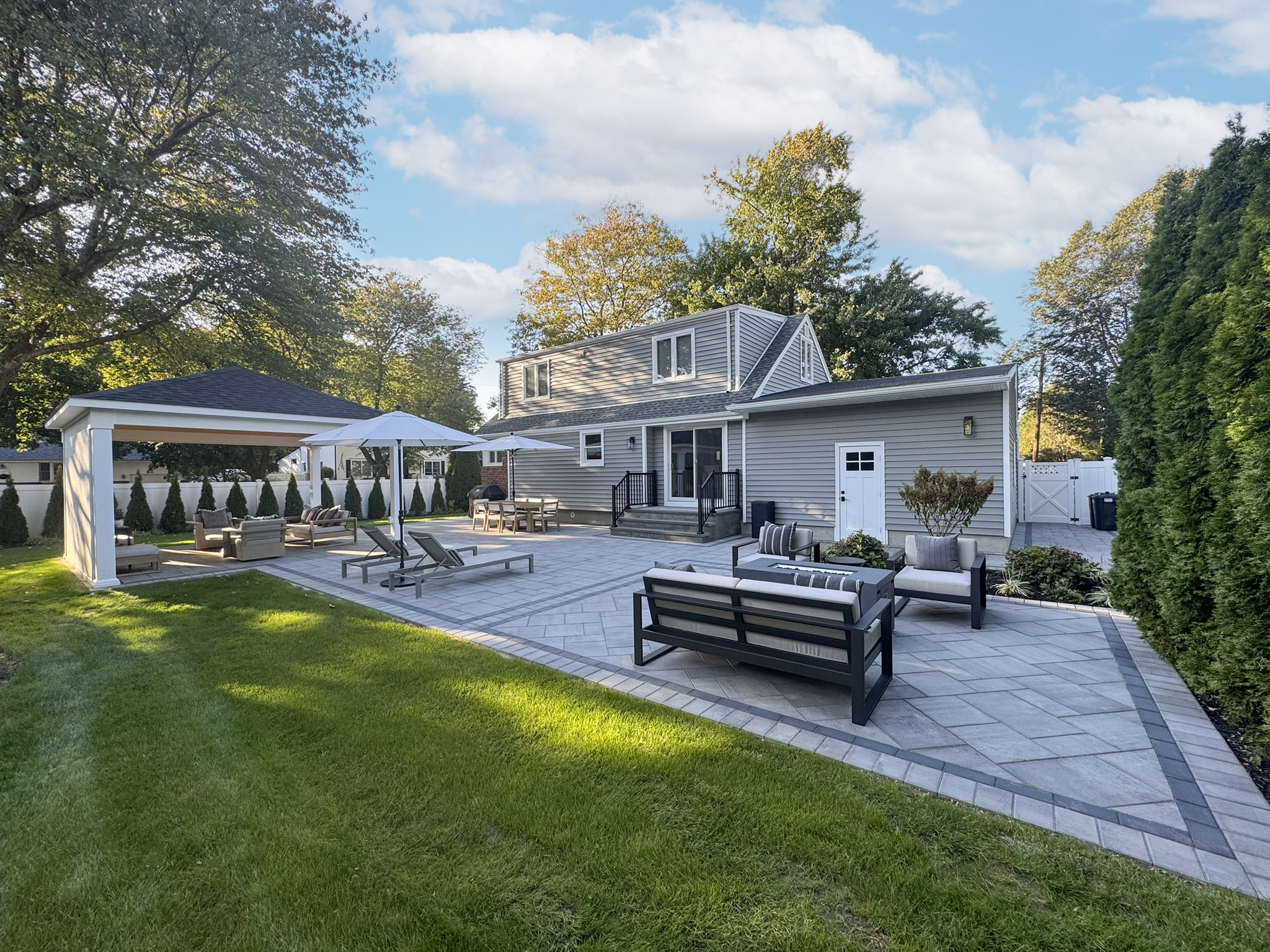 Oversized gray paver patio with contrasting soldier course border, modular outdoor sofa, open pergola lounge, and arborvitae privacy landscaping — Suffolk County NY