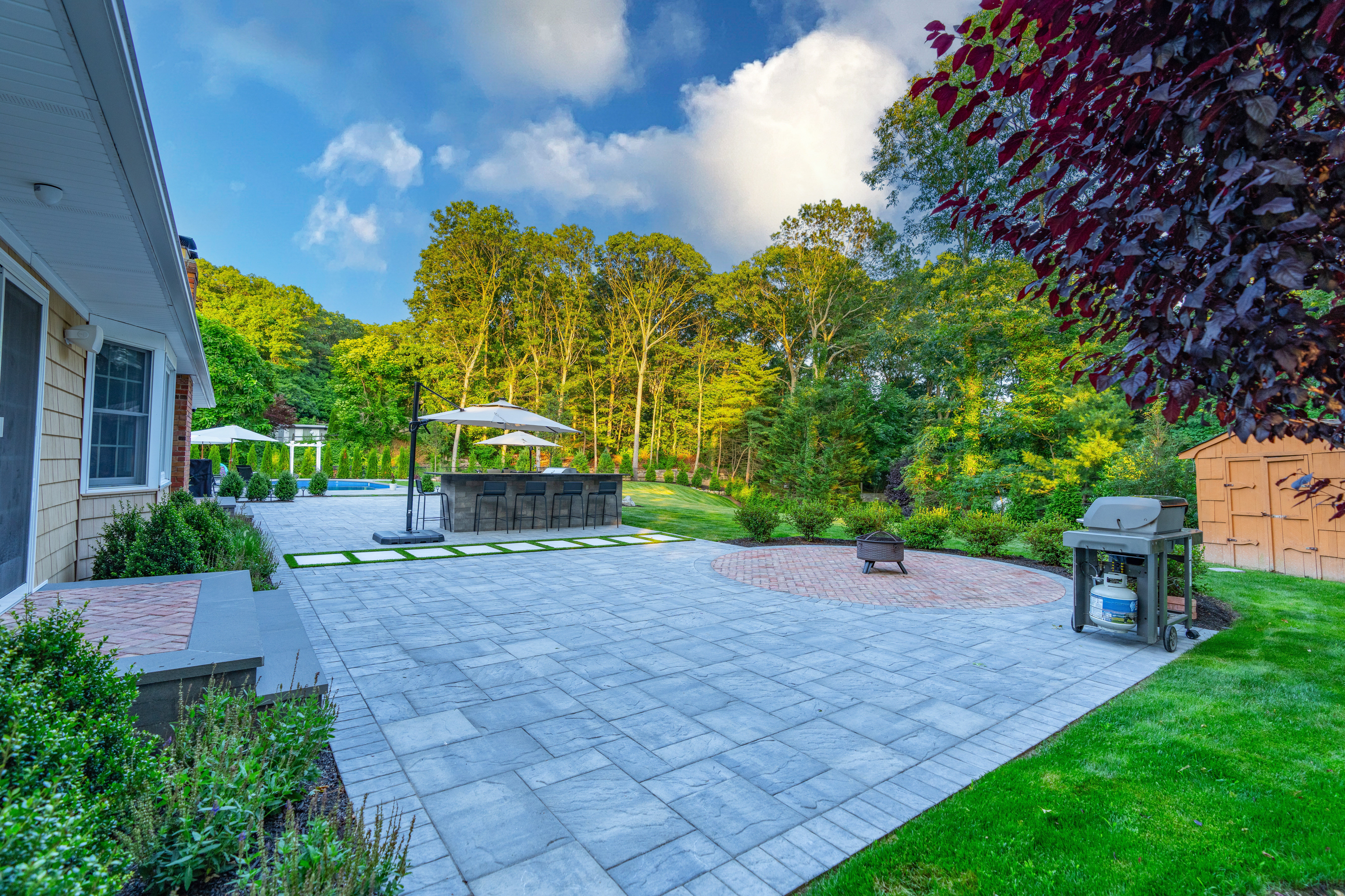 Full outdoor living transformation with large-format gray pavers, synthetic turf stepping stone path, outdoor kitchen, pergola, and inground pool — Long Island NY