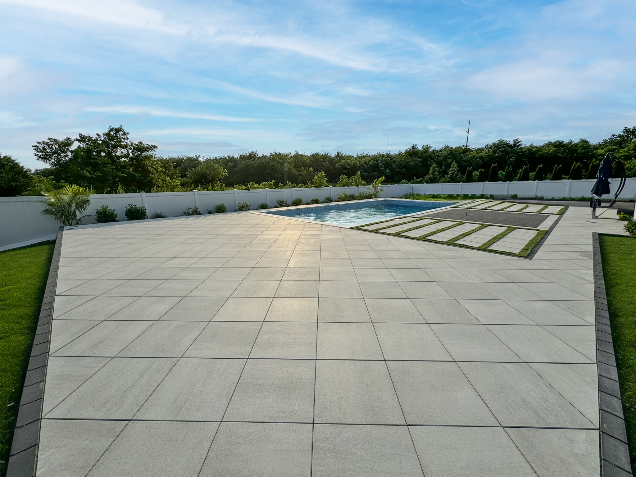 Large-format light gray porcelain paver patio with in-ground pool, synthetic turf strip walkway, and white vinyl privacy fence — Long Island NY patio contractor