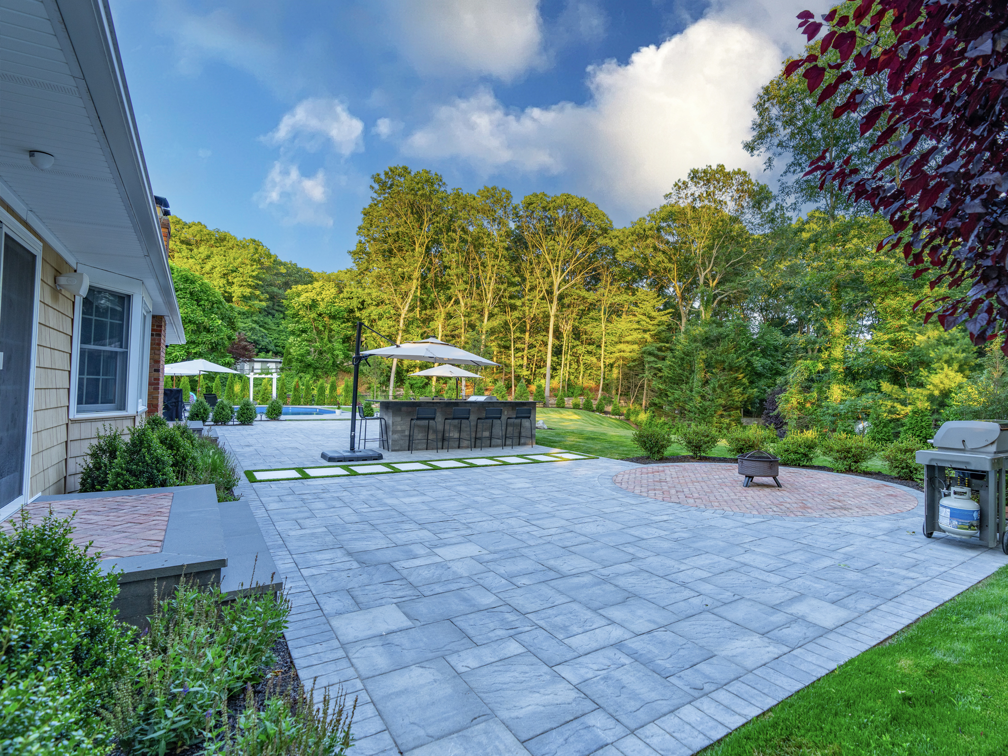 Large-format paver patio with circular brick accent area, outdoor kitchen bar, pergola, and inground pool on mature-treed Long Island property — Flawless Masonry