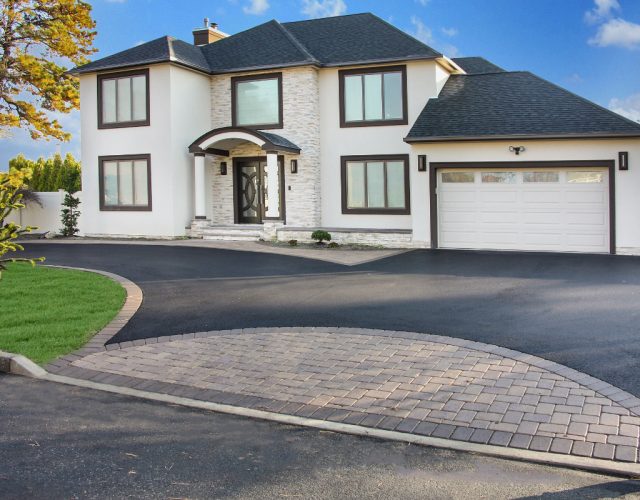 A two-story house with white exterior, dark-framed windows, and a stone entryway. It has a black asphalt driveway with a brick-bordered section in front. The garage door is white.