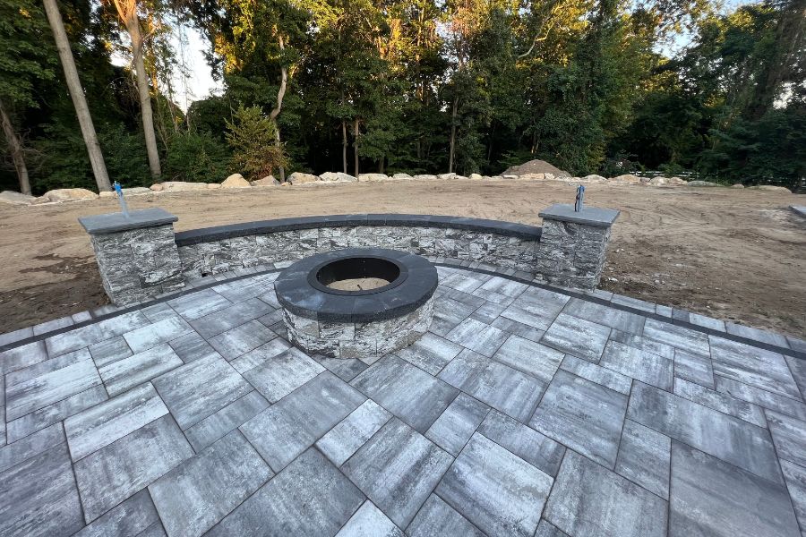 The stone patio features a circular fire pit and a curved seating wall crafted from Cambridge Pavingstones, all nestled among trees in Hauppauge, with an unfinished dirt area providing a rustic touch.