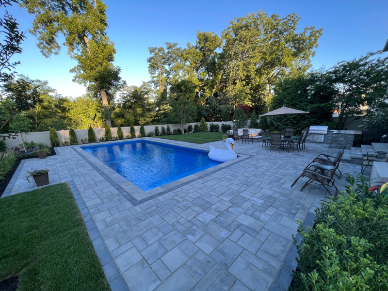 Backyard with a rectangular pool, a large inflatable swan, and surrounding patio furniture. Trees and shrubs line the fence in the background.