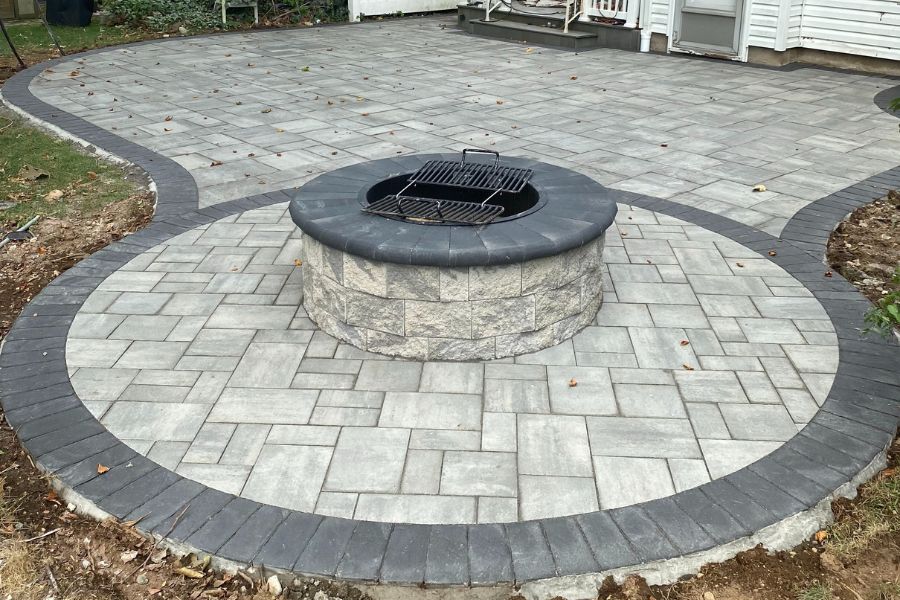 A circular stone fire pit graces the gray paved patio, bordered elegantly in dark tones. This inviting space, adjacent to a white building, offers a perfect retreat reminiscent of classic Cambridge charm.