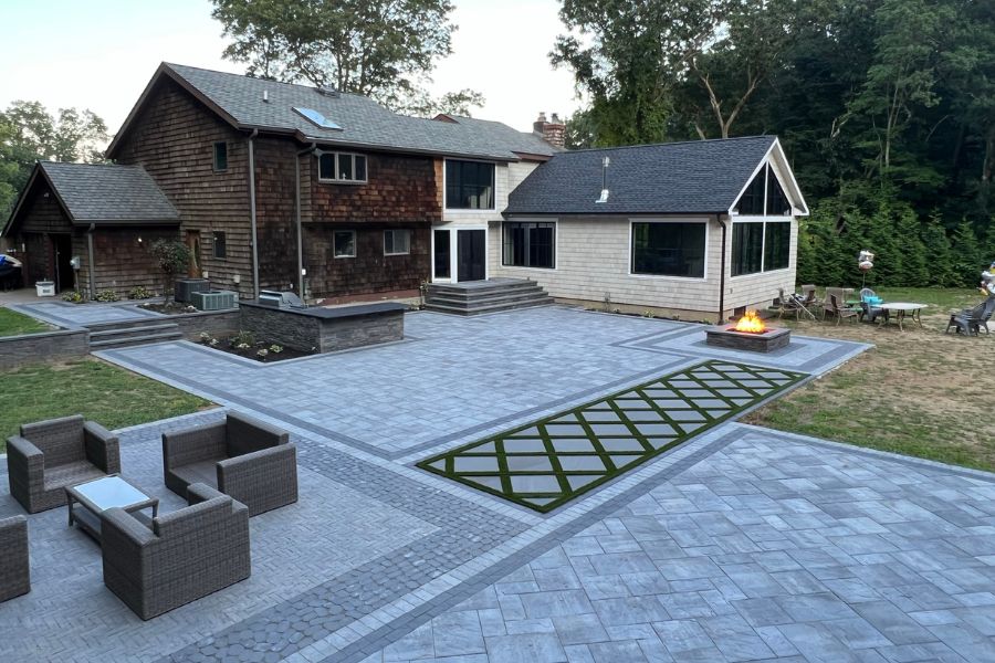 A large house in Manhasset features a stone patio with Cambridge Pavingstones, outdoor seating, and a cozy fire pit. The landscaping includes grass squares and is beautifully surrounded by trees.