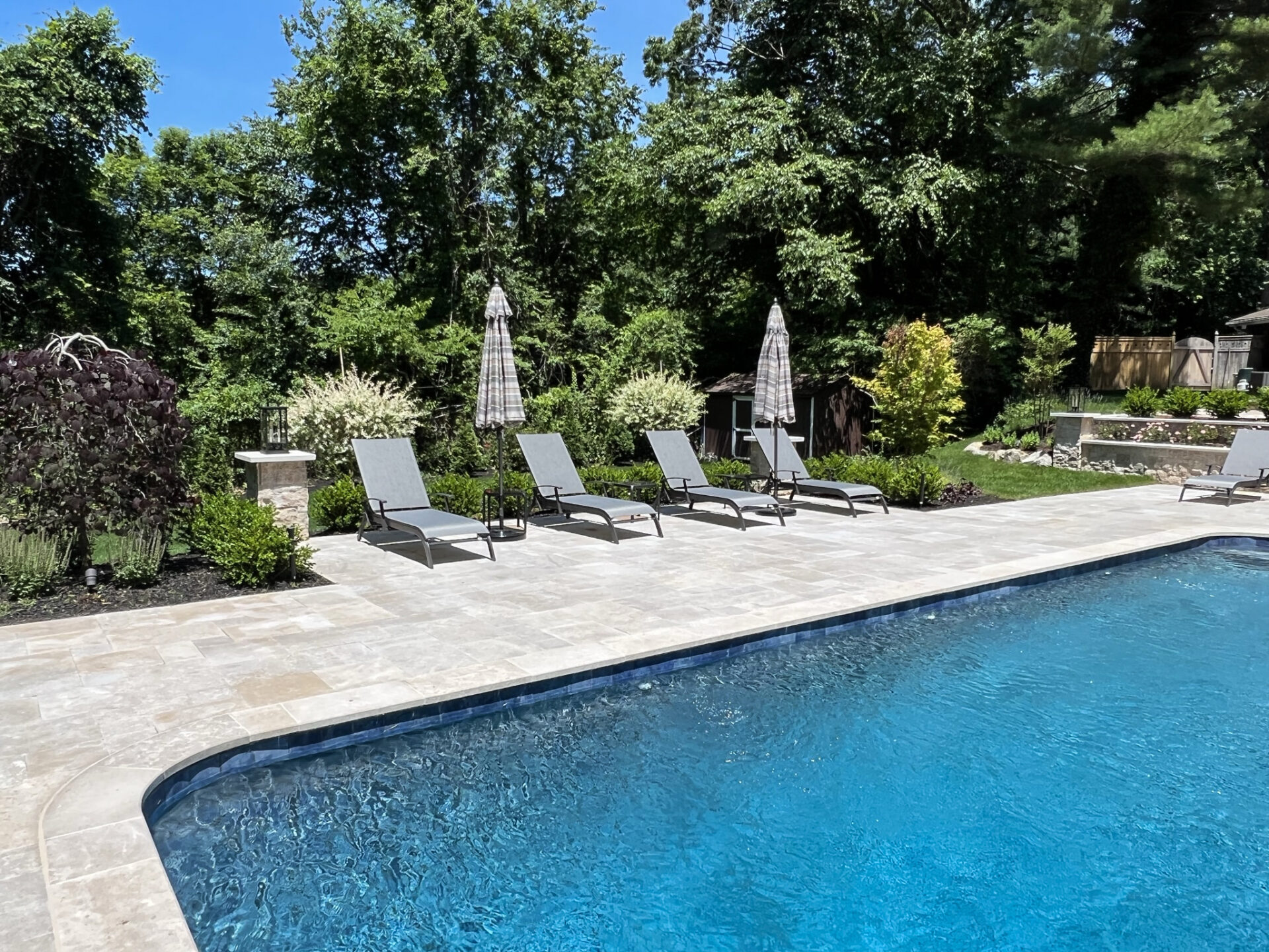 Three lounge chairs and two umbrellas are arranged on a stone pool deck beside a clear blue pool, with trees and greenery in the background.