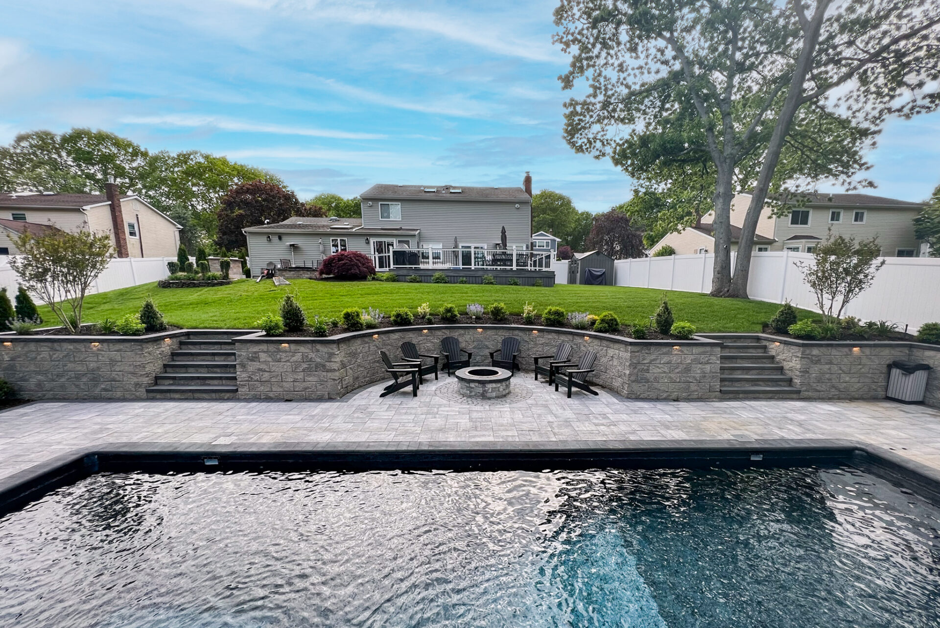 Backyard with swimming pool, stone patio, fire pit surrounded by chairs, terraced landscaping, steps, and a two-story house with deck in the background.