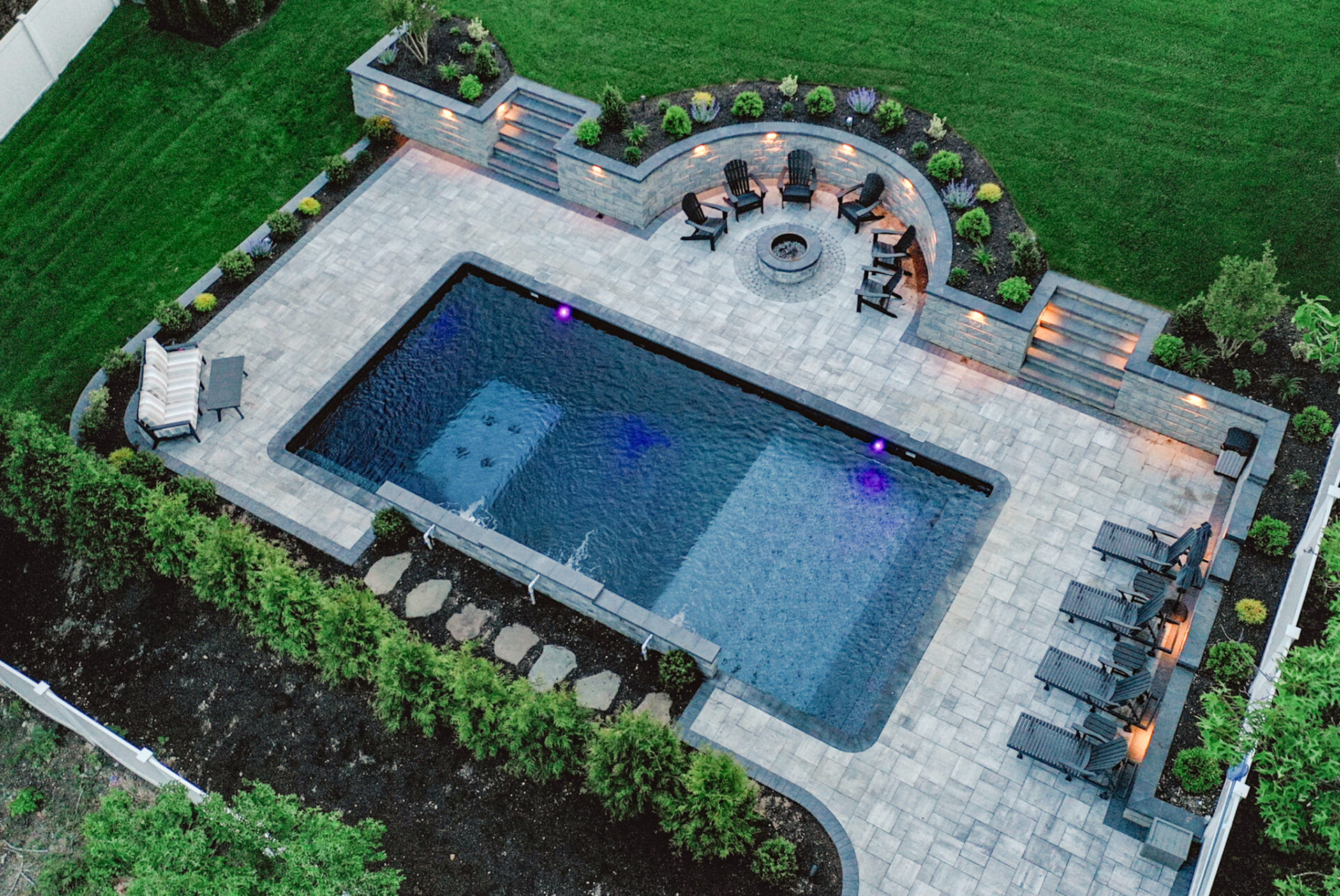 Aerial view of a rectangular backyard pool with lounge chairs, a fire pit area surrounded by chairs, stone patio, and landscaped greenery.