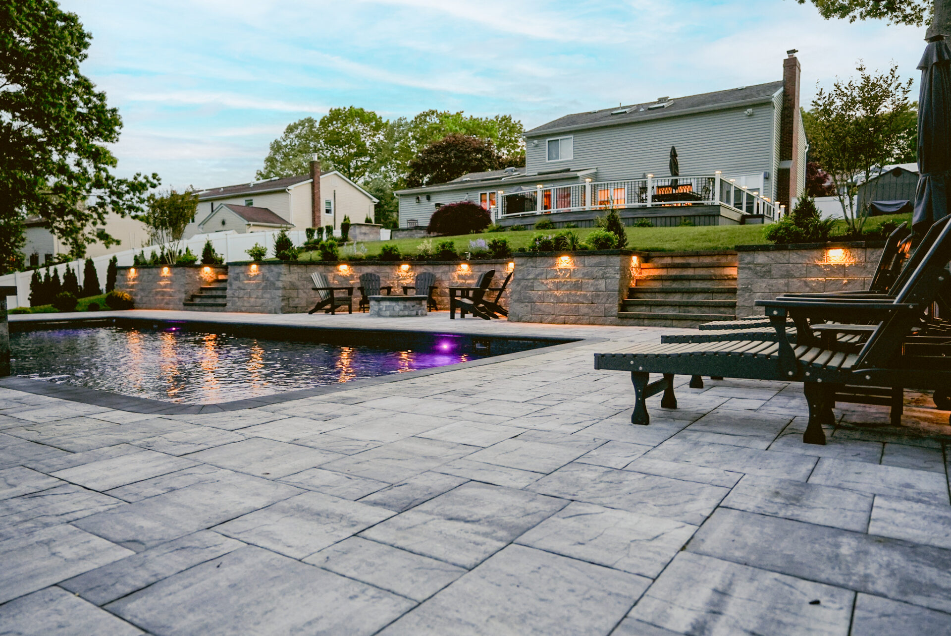 Backyard with stone patio, in-ground pool, lounge chairs, fire pit area, and raised deck behind a two-story house at dusk.