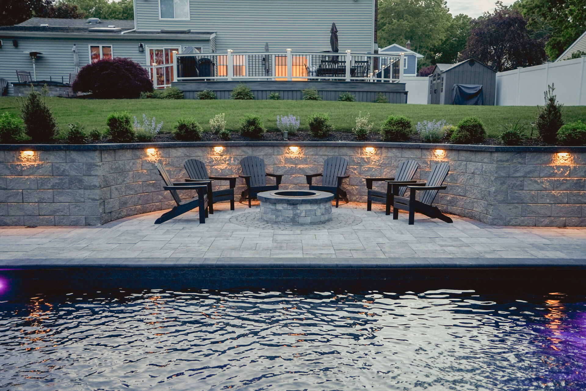 A backyard patio features Adirondack chairs arranged around a fire pit, with stone walls, outdoor lights, and a pool in the foreground.