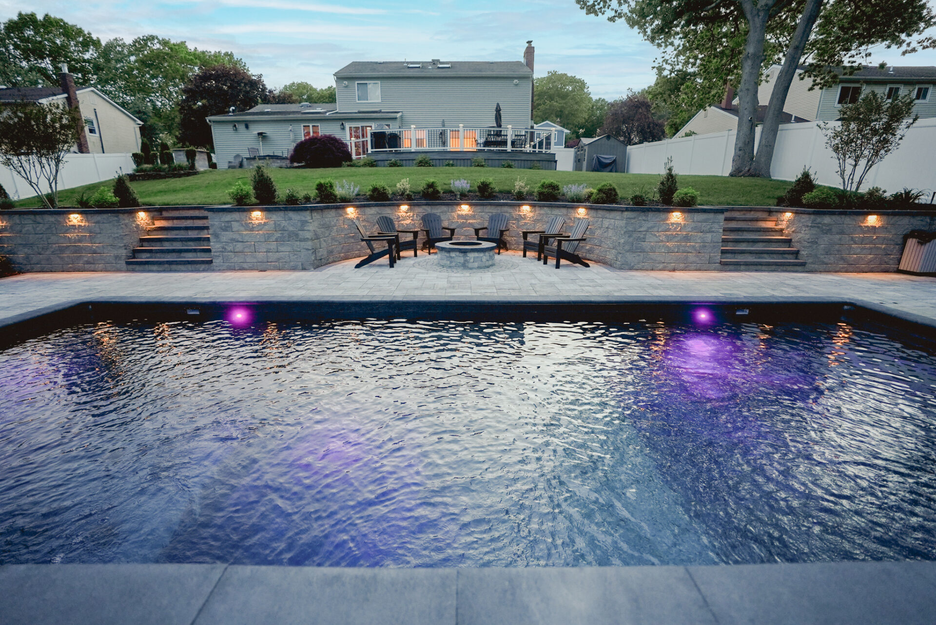 A backyard with a rectangular pool lit by purple lights, stone patio, fire pit area with chairs, and landscaped lawn surrounded by a fence and trees.