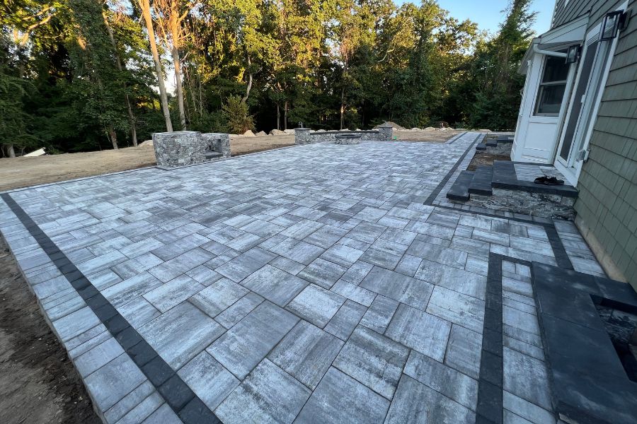 A newly constructed stone patio featuring Cambridge Pavingstones with a contrasting border sits adjacent to a house in Hauppauge, surrounded by lush trees.