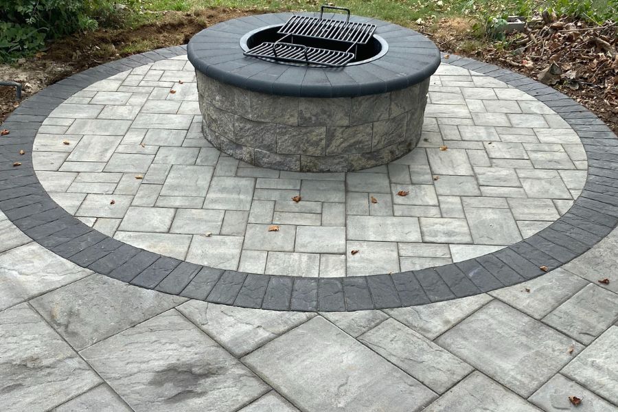 A stone fire pit, crafted with the Cambridge firepit kit, is surrounded by a circular paved patio with a contrasting Cambridge pavingstone border.