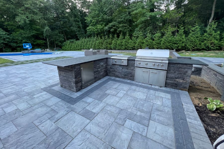 An outdoor kitchen featuring a stone countertop with a blaze grill, set on elegant pavers. A metal cabinet completes this tiled patio, surrounded by lush green hedges and trees, creating an inviting atmosphere for alfresco dining.