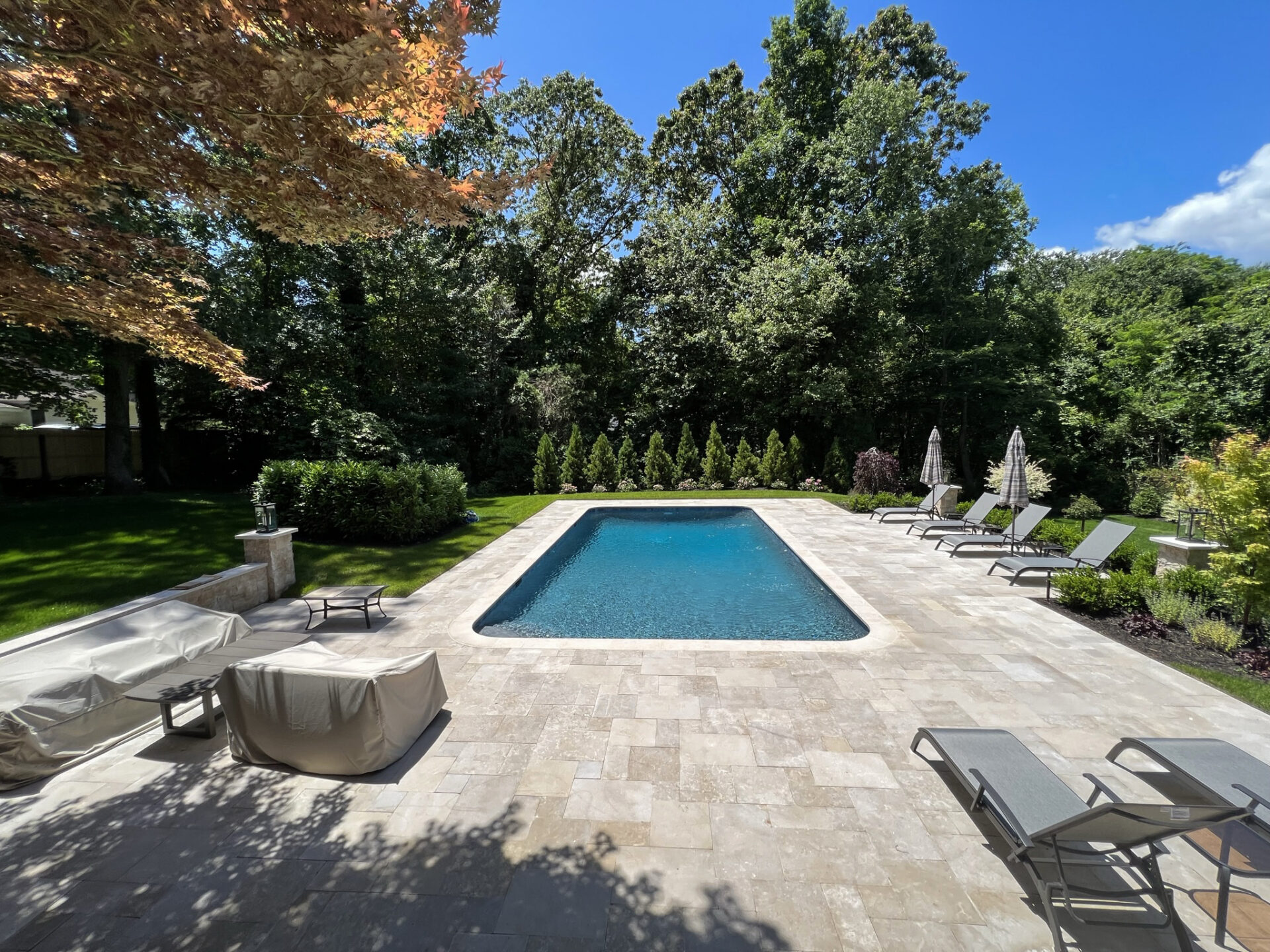 Rectangular outdoor swimming pool surrounded by lounge chairs and trees under a clear blue sky, with some furniture covered in the foreground.
