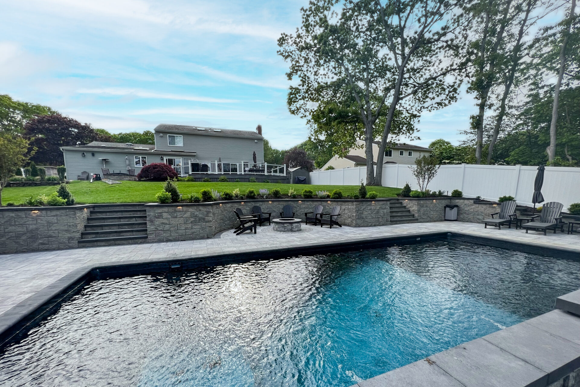 A backyard features a rectangular swimming pool, stone patio, fire pit area with seating, lawn, and a two-story gray house with a white fence and trees in the background.