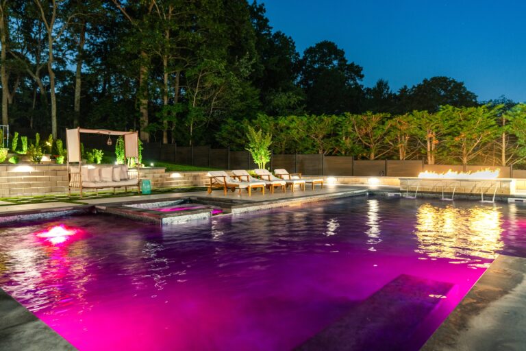 Outdoor swimming pool at night with purple lighting, lounge chairs, a fire feature, greenery, and seating area in the background.