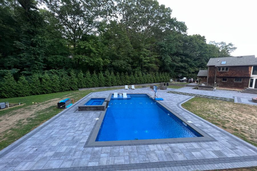 A rectangular pool with a small adjacent hot tub sits in a charming backyard surrounded by trees and a cozy wooden house. The pool patio is elegantly paved with gray stones, creating an inviting oasis that's reminiscent of Long Island's serene landscapes.