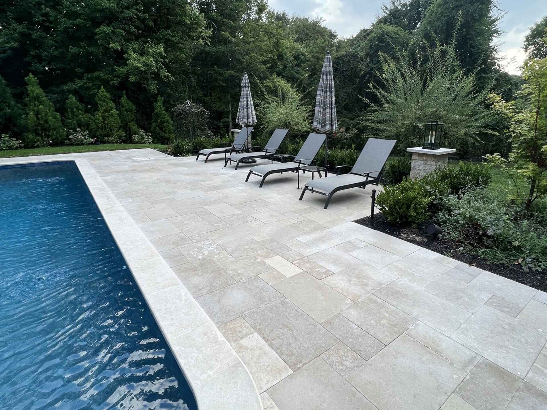 Four gray lounge chairs with two closed umbrellas sit on a stone pool deck beside a rectangular pool, surrounded by greenery and trees.