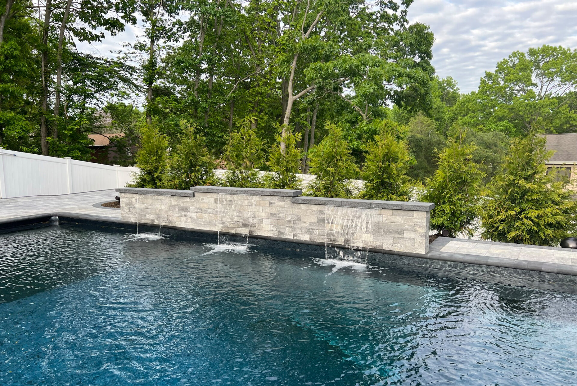 Rectangular outdoor swimming pool with water features flowing from a stone wall, surrounded by trees and greenery.