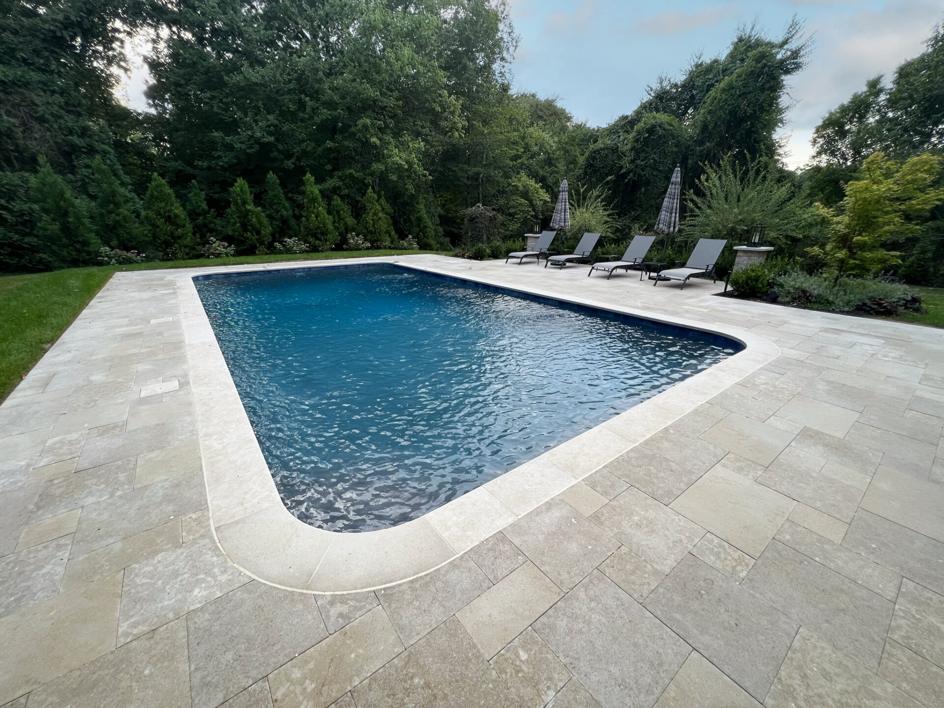 Rectangular outdoor swimming pool with stone tile deck, surrounded by trees and bushes, with several lounge chairs and umbrellas along one side.
