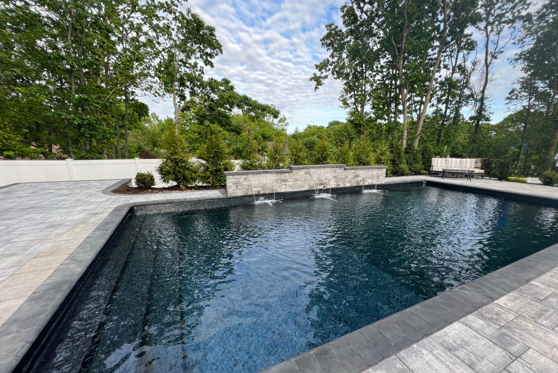 Rectangular backyard pool with three fountain spouts, surrounded by stone tiles, trees, and a white fence in the background.