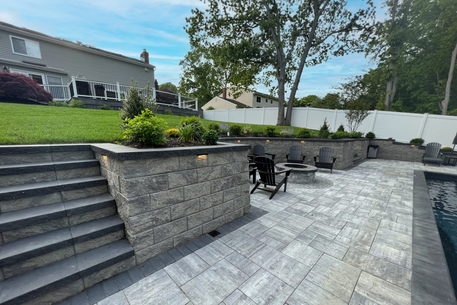 A backyard with stone steps, a retaining wall, patio seating around a fire pit, landscaped greenery, and a swimming pool.