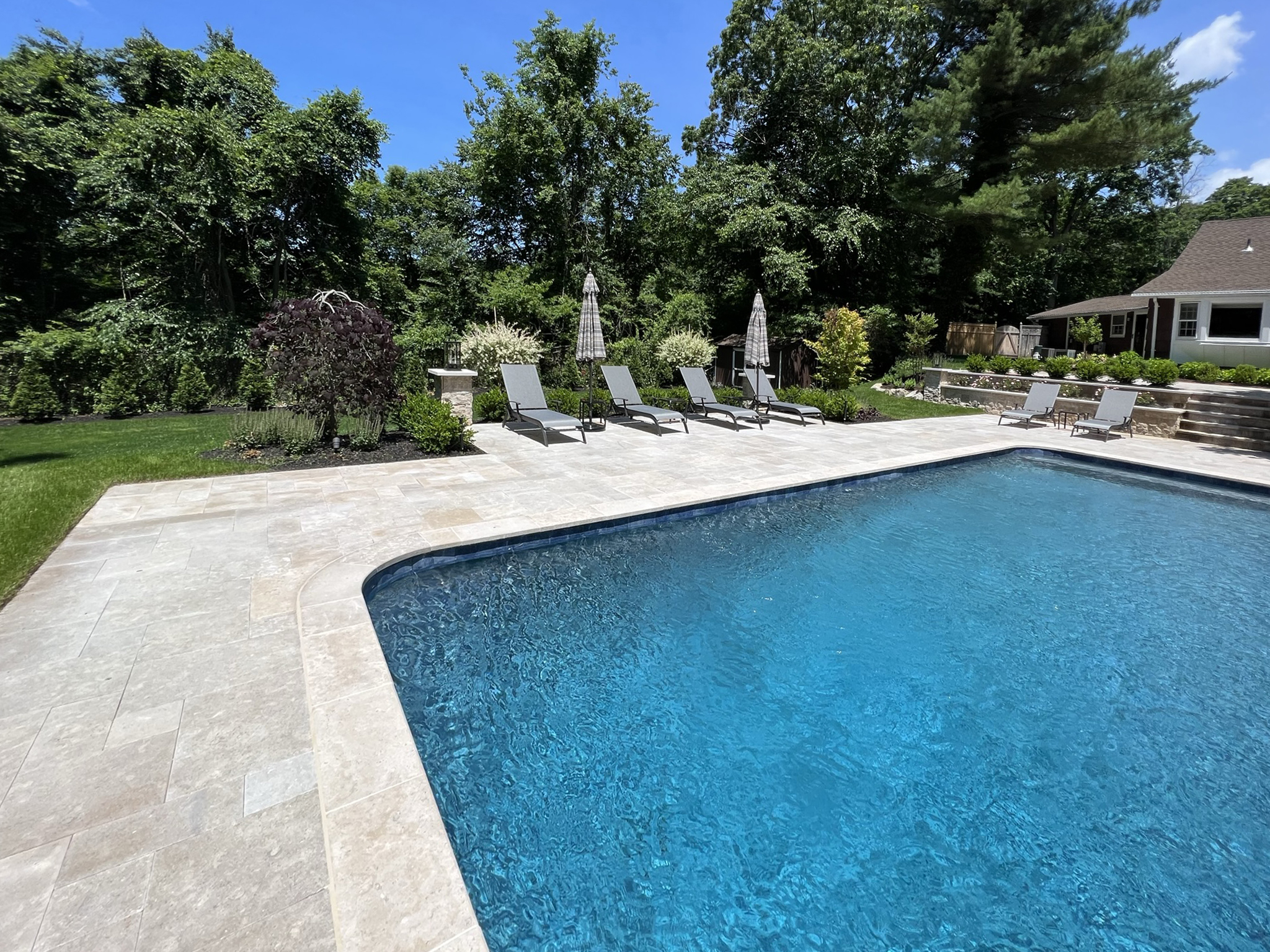 A backyard swimming pool with clear blue water, surrounded by lounge chairs, umbrellas, green lawn, and trees under a sunny sky.