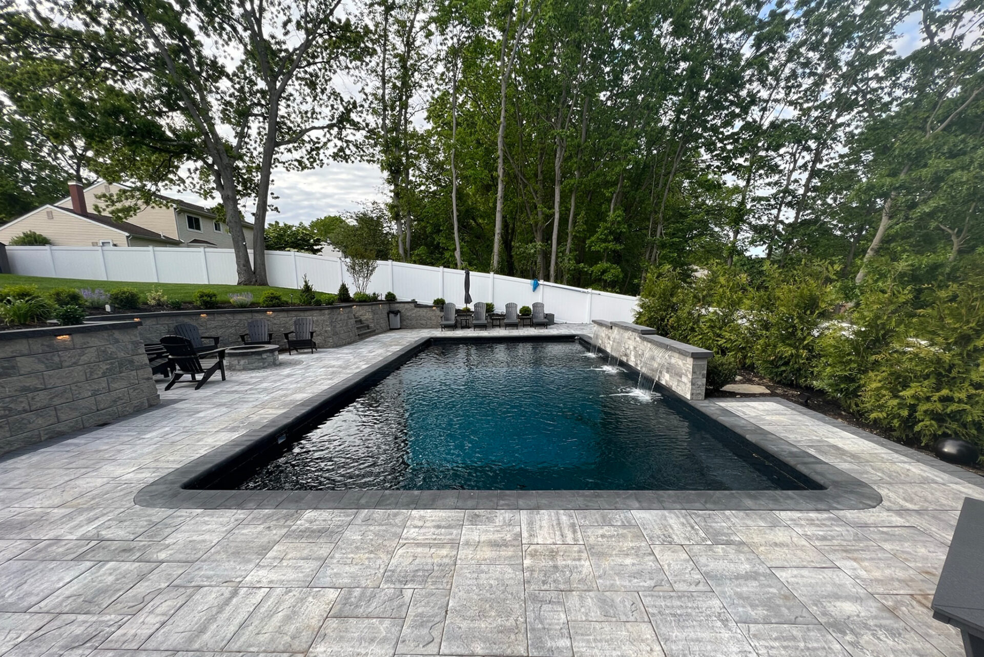 Rectangular in-ground pool with a stone tile deck, black lounge chairs, and water features, surrounded by trees and privacy fencing.