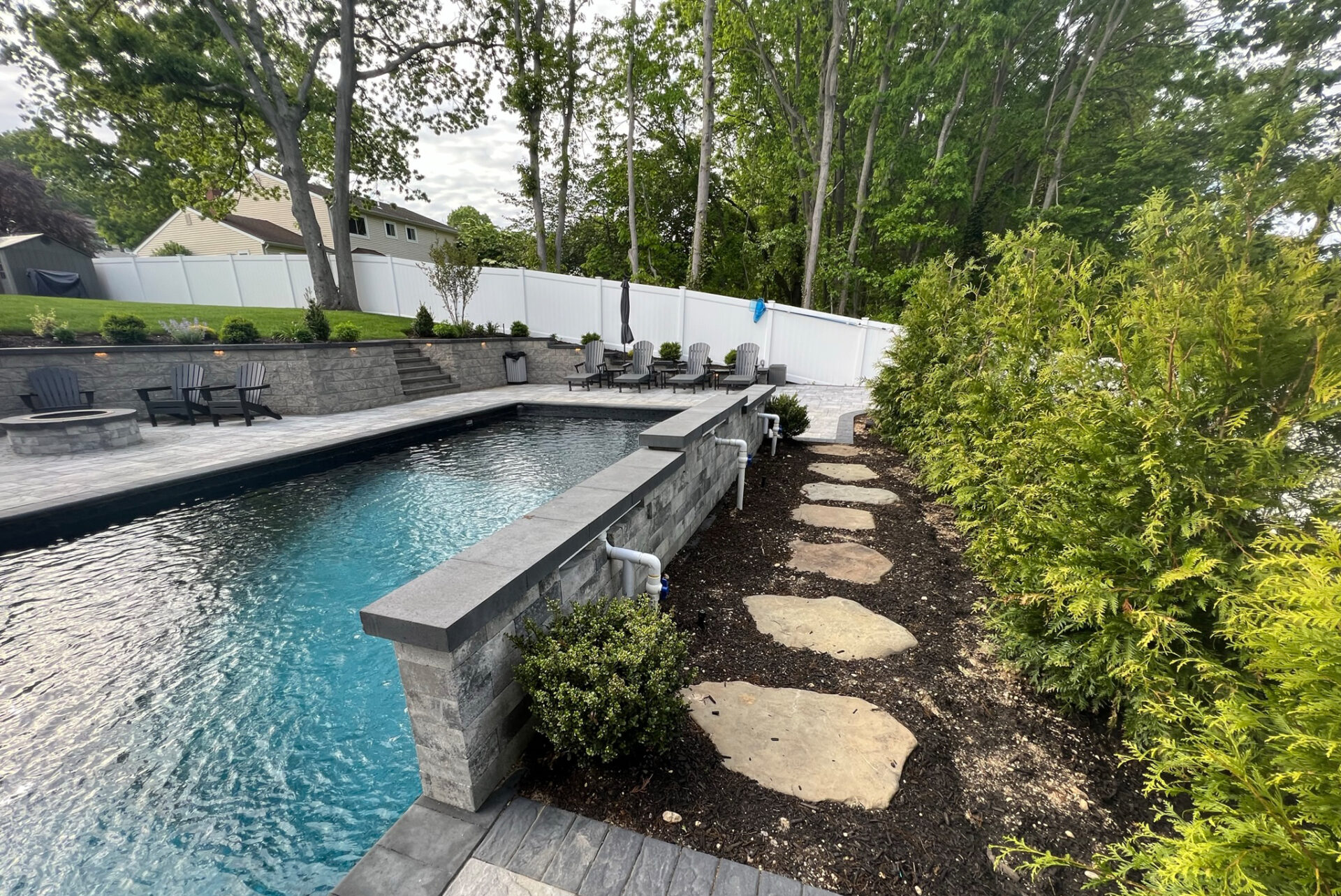 A backyard with a rectangular swimming pool, stone walkway, lounge chairs, small shrubs, and tall trees along a white privacy fence.