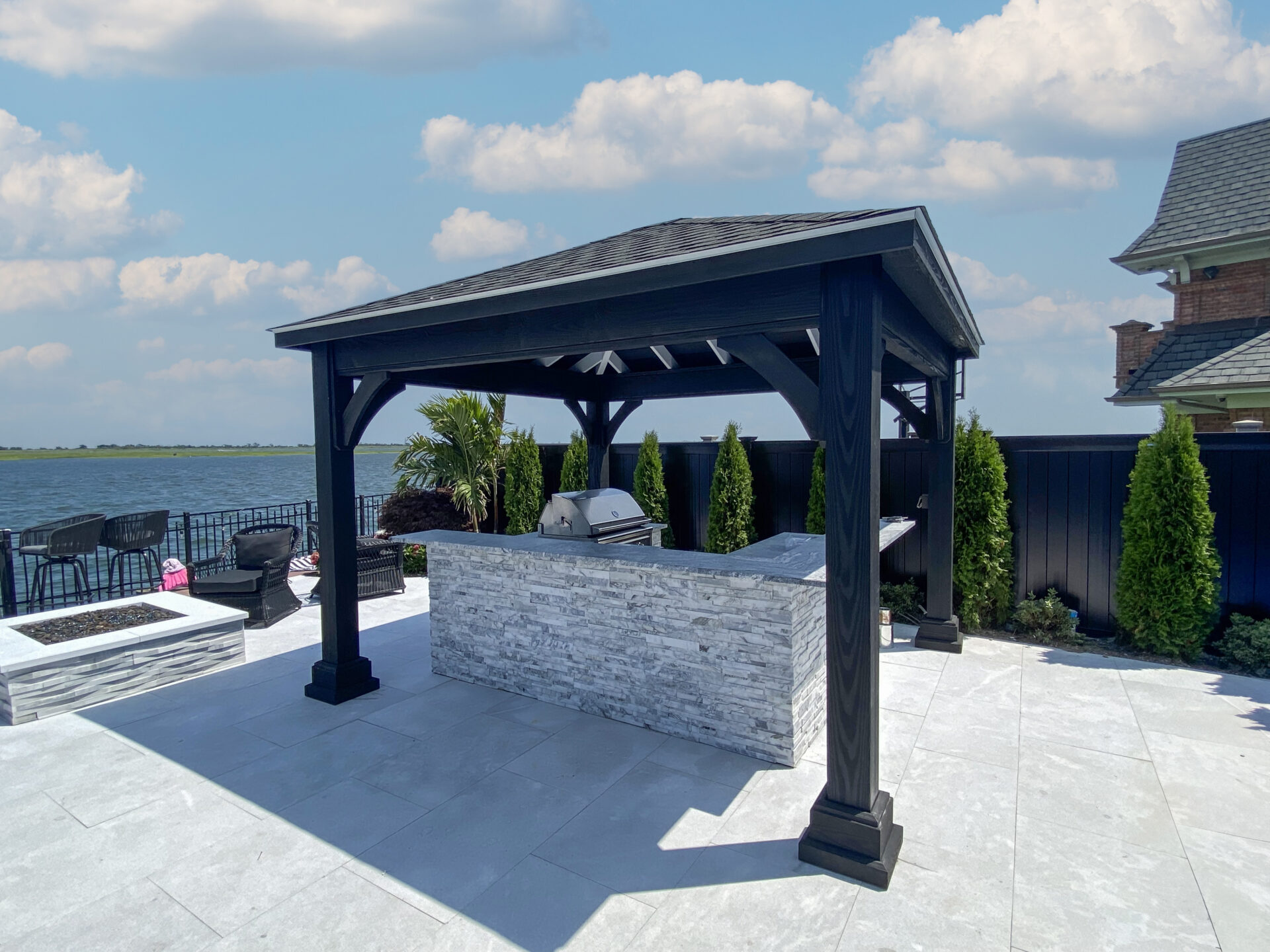 A modern outdoor kitchen with a stone counter and grill under a black pavilion, set on a patio by the water with greenery and seating nearby.