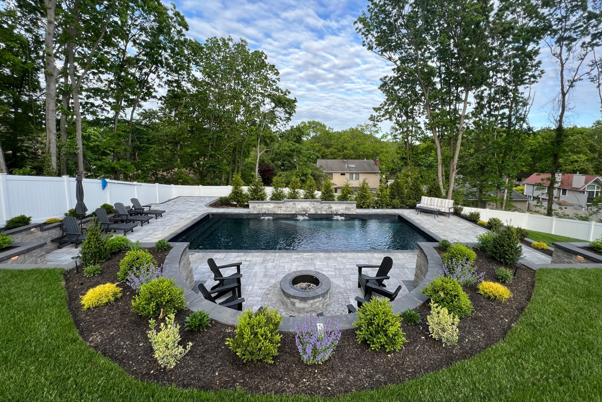 Backyard with an in-ground pool, stone patio, fire pit with chairs, landscaped garden beds, and surrounding trees under a partly cloudy sky.