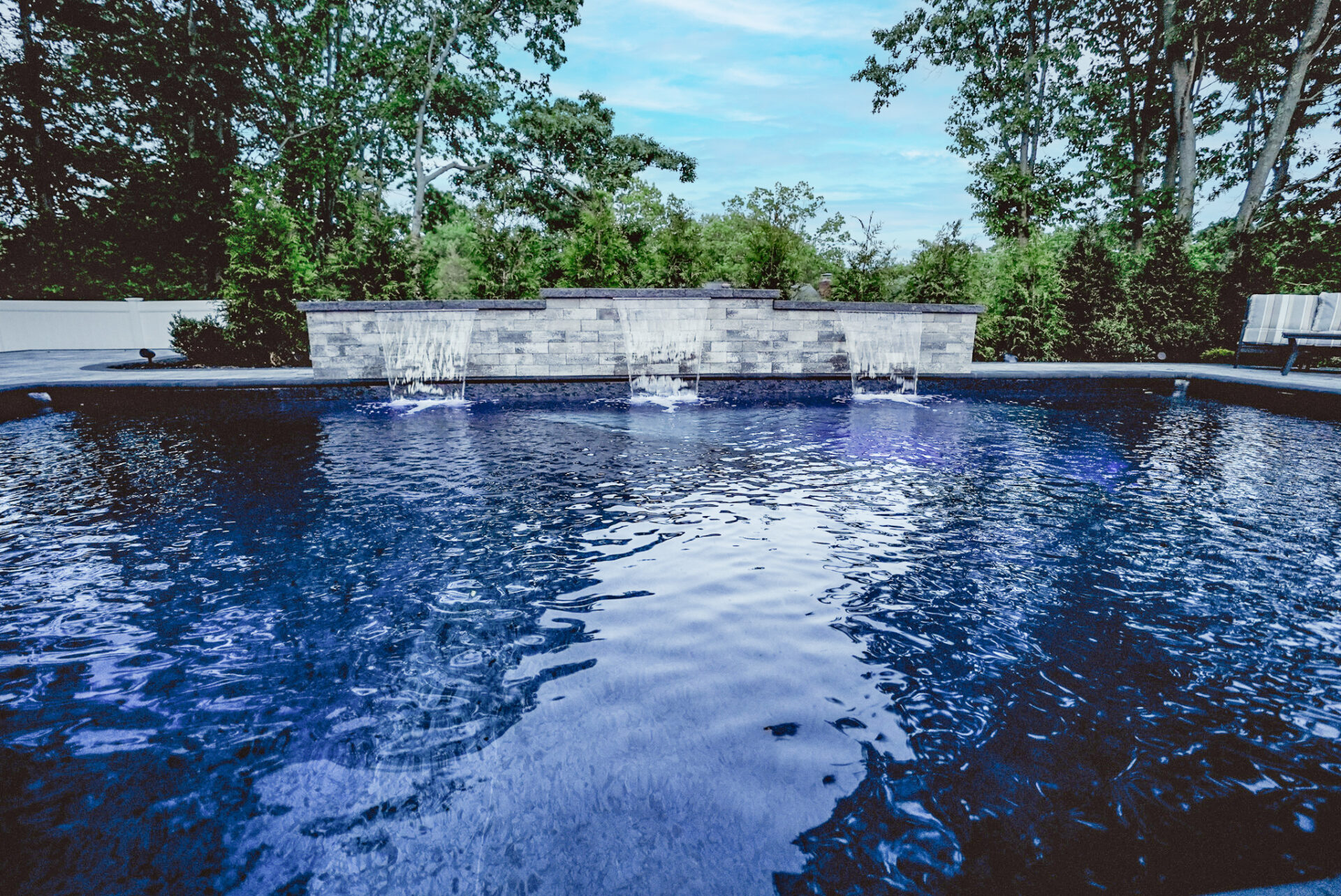 A backyard swimming pool with four small waterfalls flowing from a stone wall, surrounded by trees and greenery.