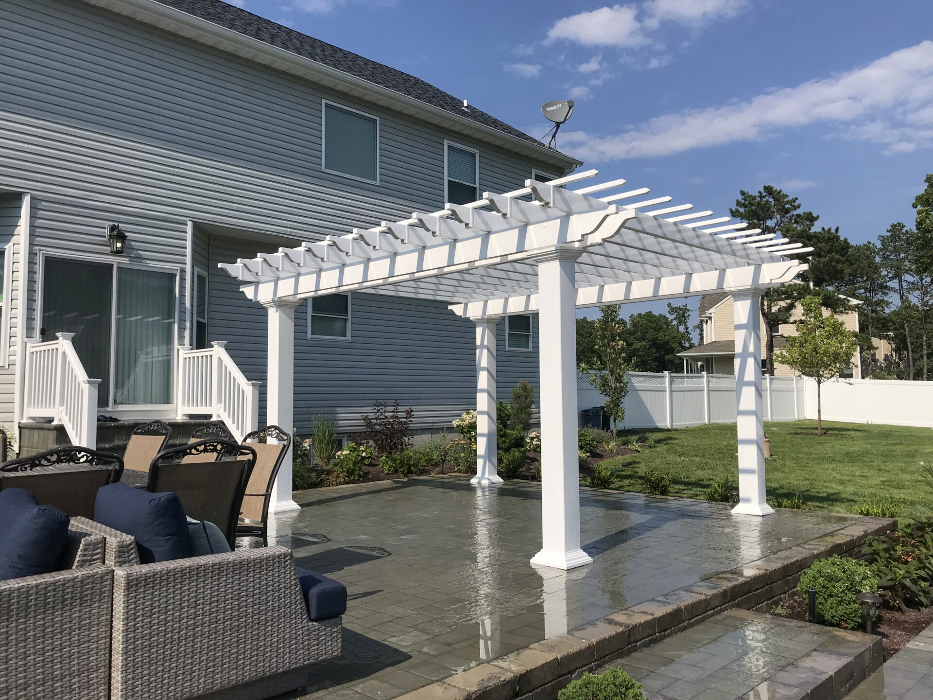 A white pergola stands on a paved patio in a backyard, near wicker outdoor furniture and behind a two-story gray house.