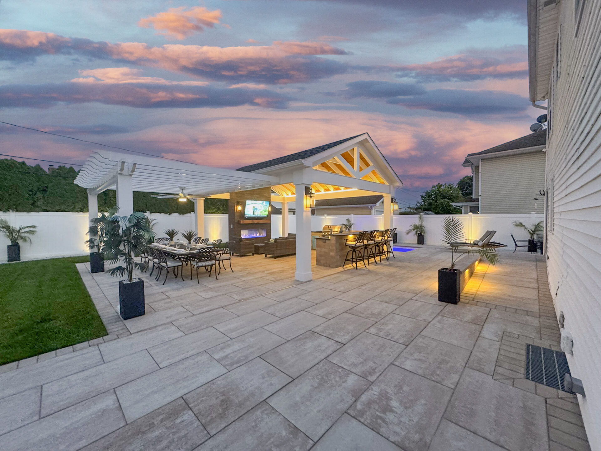 Outdoor patio with covered seating, bar area, dining table, potted plants, and built-in lighting at sunset; modern design with tiled flooring and white privacy walls.