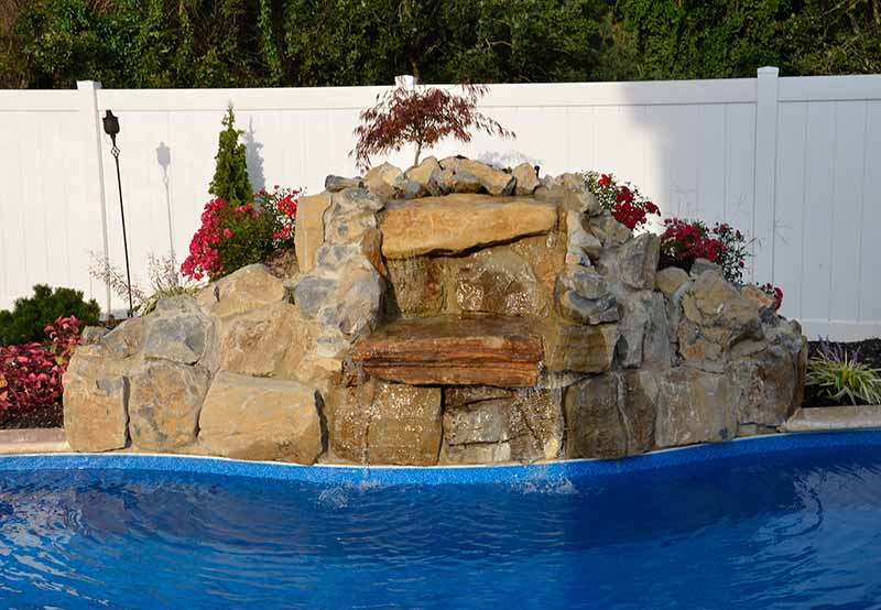 Artificial rock waterfall by a swimming pool, surrounded by lush plants and white fence.