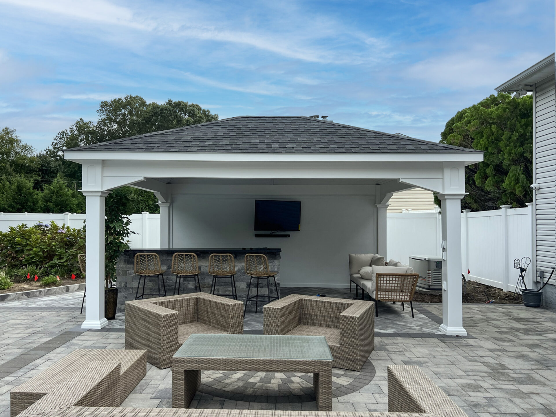 A covered outdoor patio with a TV, bar stools, wicker seating, and a coffee table set on a stone-paved area beside a white fence and house.