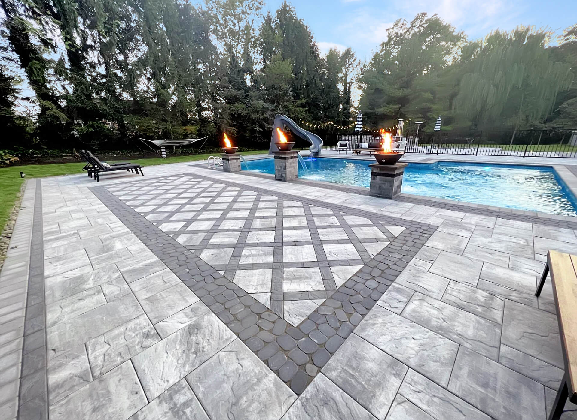A backyard with an in-ground pool, poolside fire pits, lounge chairs, a hammock, and surrounding trees. The area is paved with patterned stone tiles.