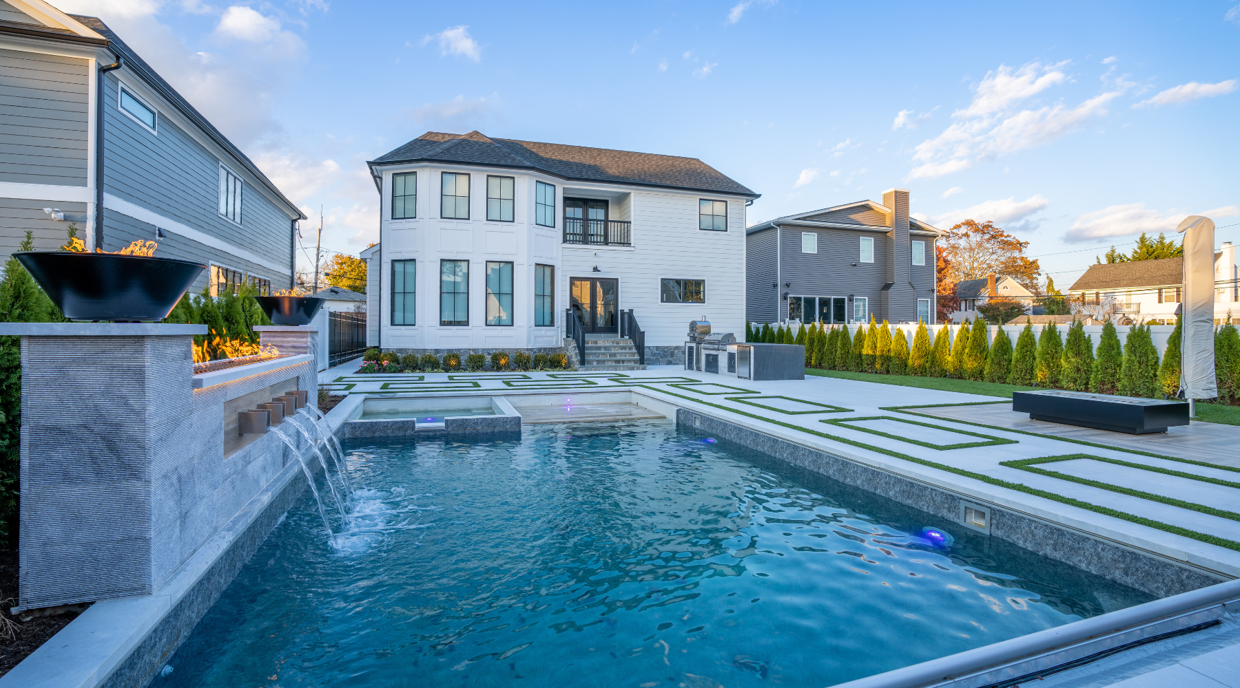 Backyard view with a large modern rectangular pool, geometric patio, and a fire feature. A white two-story house and surrounding greenery are visible in the background under a partly cloudy sky.