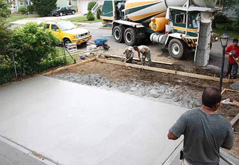 Workers pour and level concrete from a mixer truck for a new driveway, with tools and vehicles nearby.