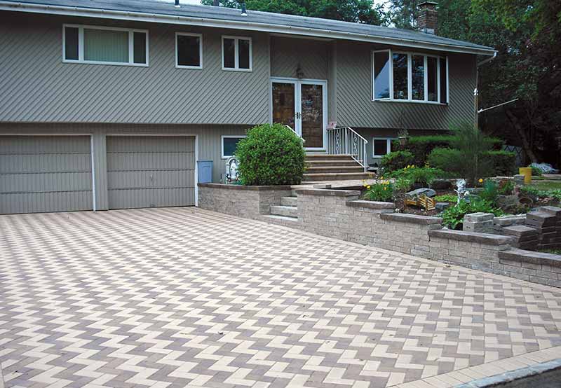A split-level house with a two-car garage and a brick driveway featuring a herringbone pattern, surrounded by a landscaped garden.
