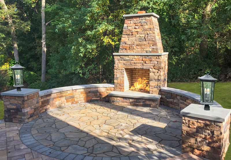A stone outdoor fireplace with a lit fire, flanked by two lanterns, set on a curved, paved patio surrounded by lush green trees.