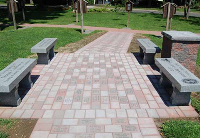 Brick pathway lined with personalized engraved bricks, flanked by benches with plaques, in a lush green park.