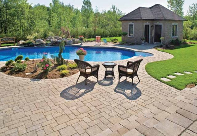 Lush backyard featuring a cobblestone patio with two chairs and a table, a large swimming pool, and a small house surrounded by greenery and bright flowers.
