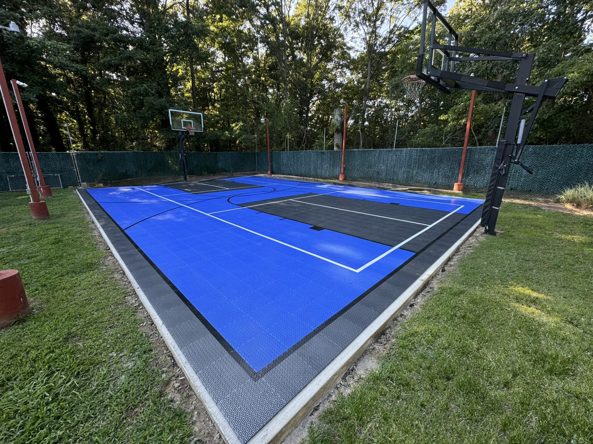Outdoor basketball court with blue and black surface, two hoops, surrounded by grass and trees, and enclosed by a green chain-link fence.