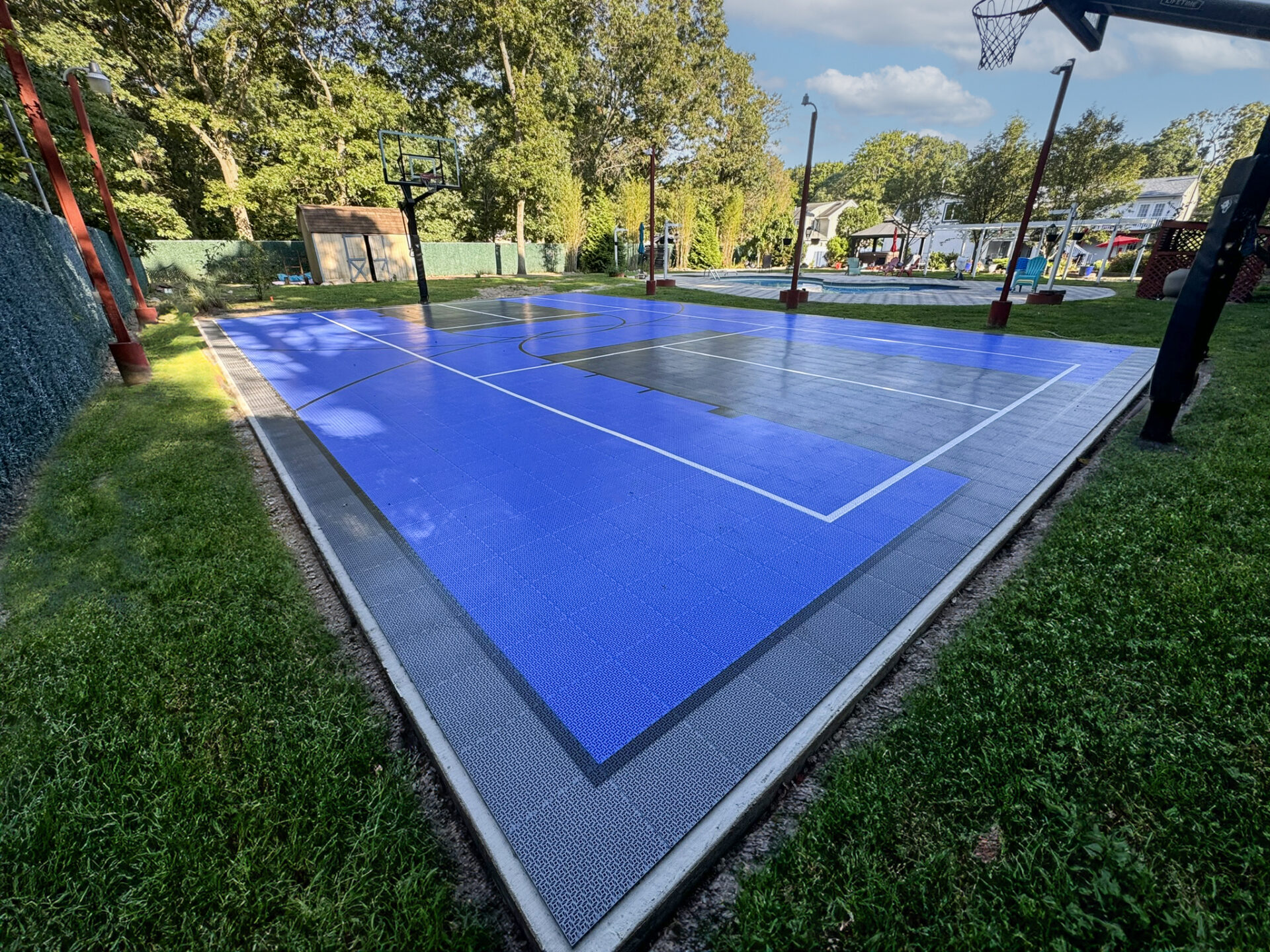Outdoor basketball court with blue and gray surface, surrounded by grass and trees, with residential houses visible in the background.