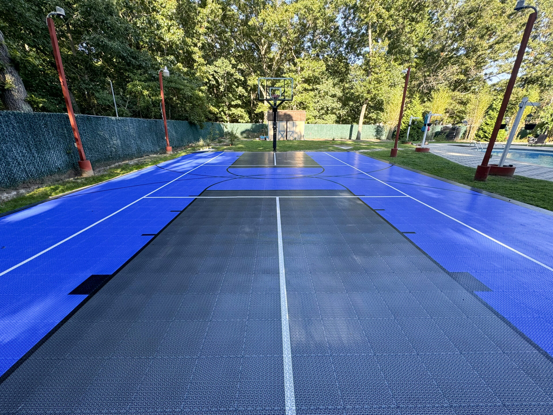 Outdoor basketball court with blue and black flooring, surrounded by a green fence and red light poles, trees in the background.