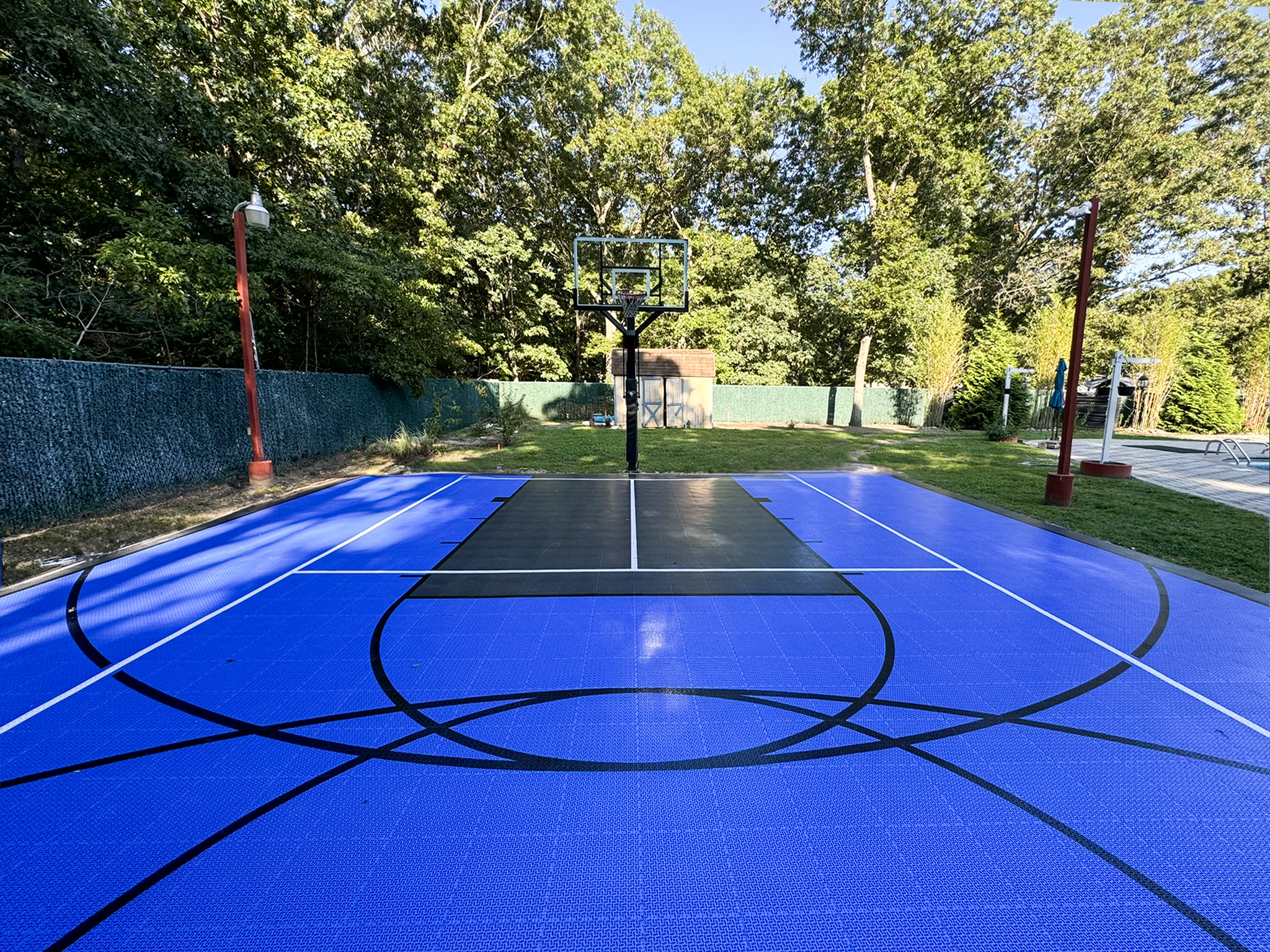 Outdoor basketball court with a blue playing surface, black key area, white boundary lines, basketball hoop, and surrounded by trees and fencing.