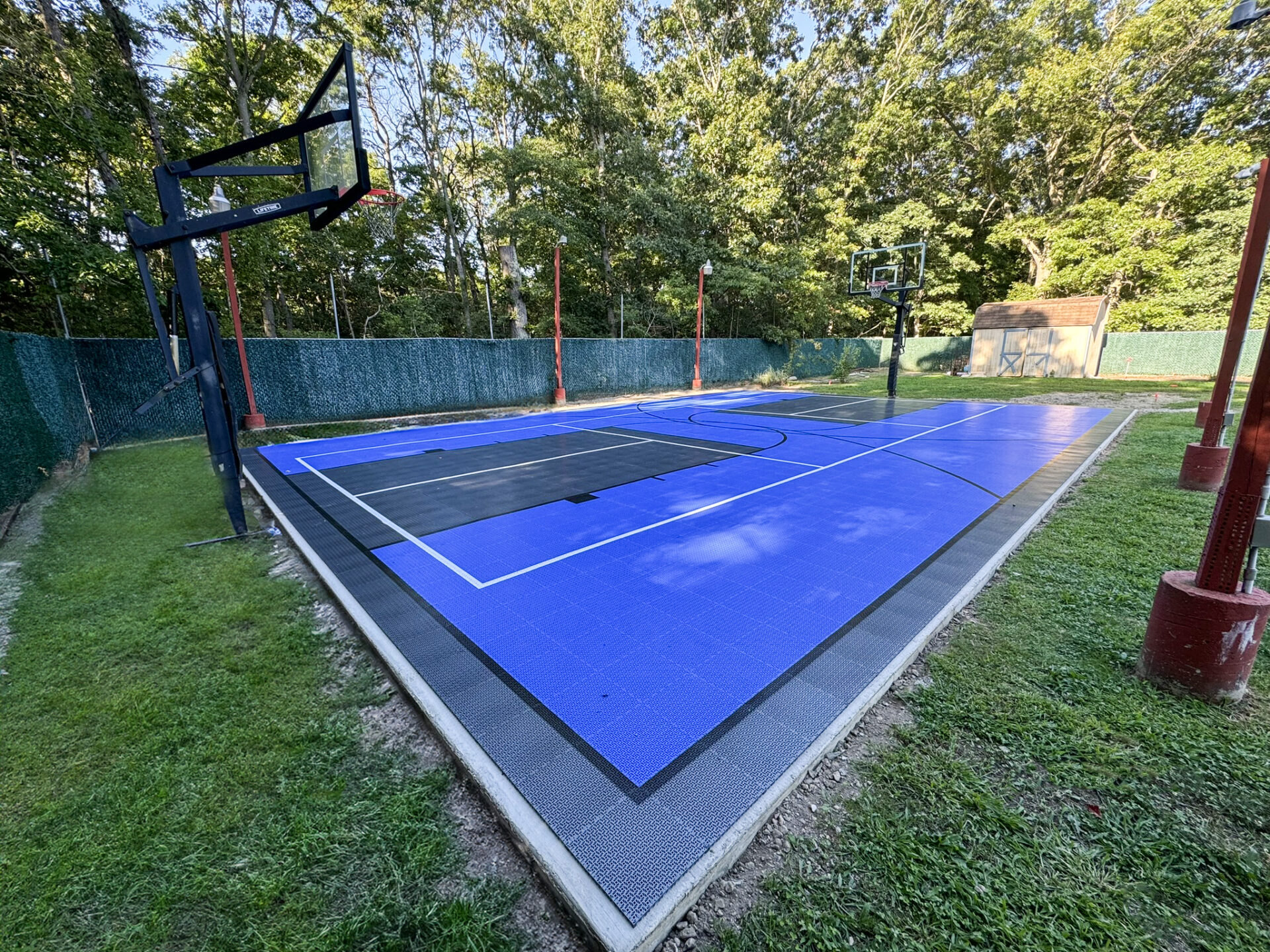 Outdoor basketball court with blue and black surface, surrounded by green fencing and trees, with two basketball hoops visible, one on each end.