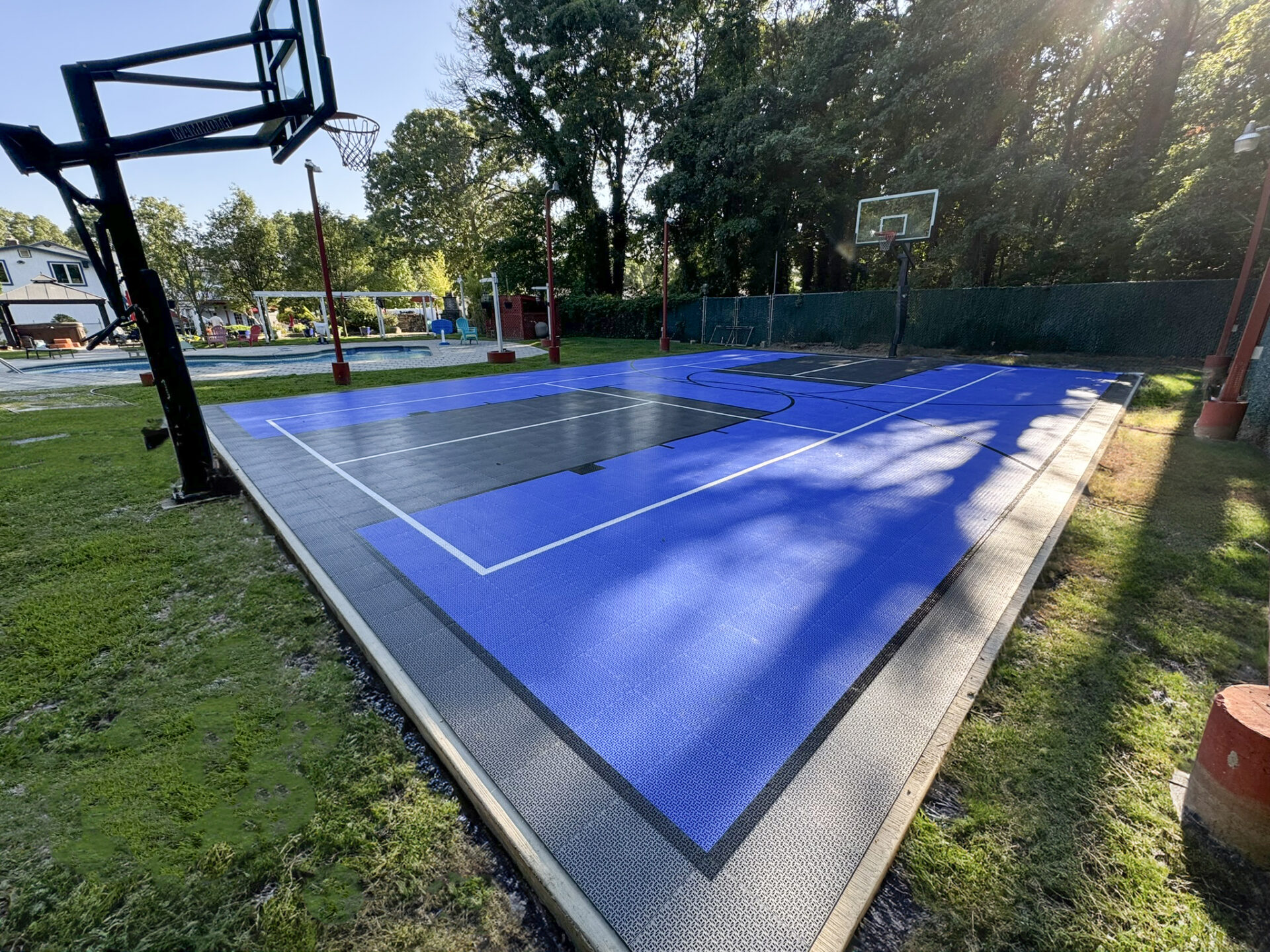 Outdoor basketball court with blue and black flooring, white boundary lines, and two hoops, surrounded by grass, trees, and a playground in the background.
